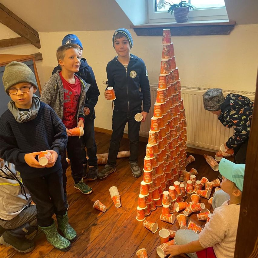 Mehrere Kinder stehen um eine Pyramide aus orangefarbenen Bechern herum, einige halten Becher, andere sind gefallen. Ein Kind sitzt auf dem Boden und repariert die Pyramide. Eine Pflanze steht auf der Fensterbank.