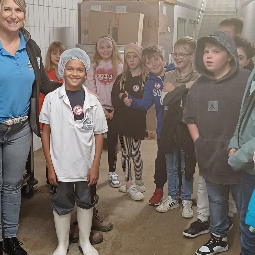 Eine Gruppe von Kindern und eine erwachsene Frau stehen in einem Flur, wahrscheinlich auf einer Schulfahrt. Die Kinder tragen verschiedene Kleidungsstücke und Schuhe. Die erwachsene Frau trägt eine Kochmütze und lächelt.