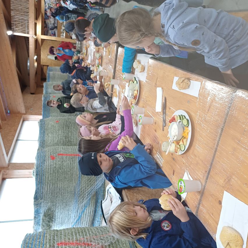 Mehrere Kinder sitzen an einem langen Tisch und essen eine Mahlzeit. Der Tisch ist mit Tellern, Servietten und Getränken gefüllt. Einige Kinder tragen Jacken, andere Mützen. In der Nähe des Tisches stehen einige Erwachsene.