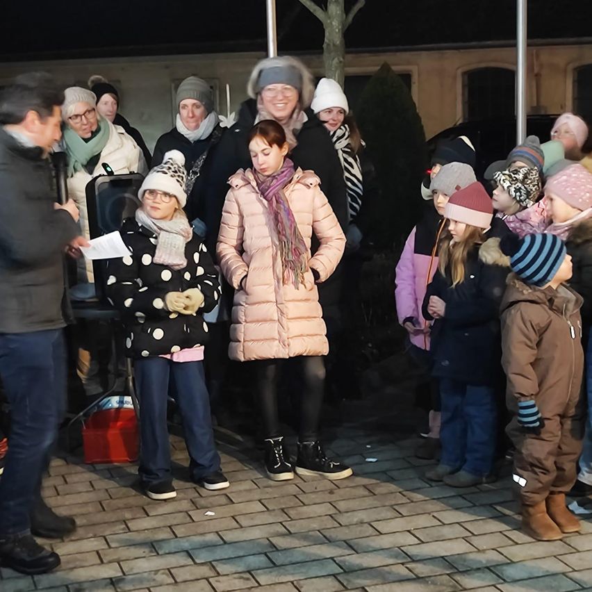 A group of adults and children dressed in winter clothing are gathered outdoors at night. A woman in a pink coat and scarf stands out, while others wear hats and gloves.