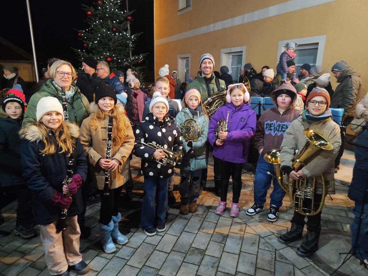 A group of children and adults with musical instruments, including trumpets and a clarinet, are standing together in front of a decorated Christmas tree.