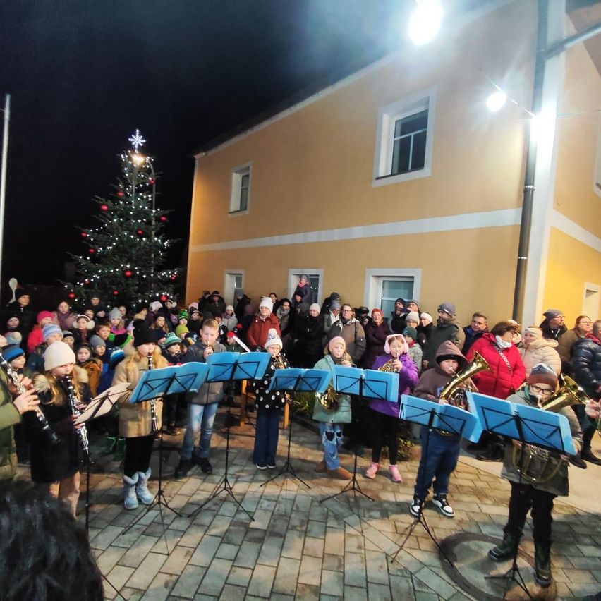 A group of children and adults are playing musical instruments in front of a decorated Christmas tree outside a yellow building at night.