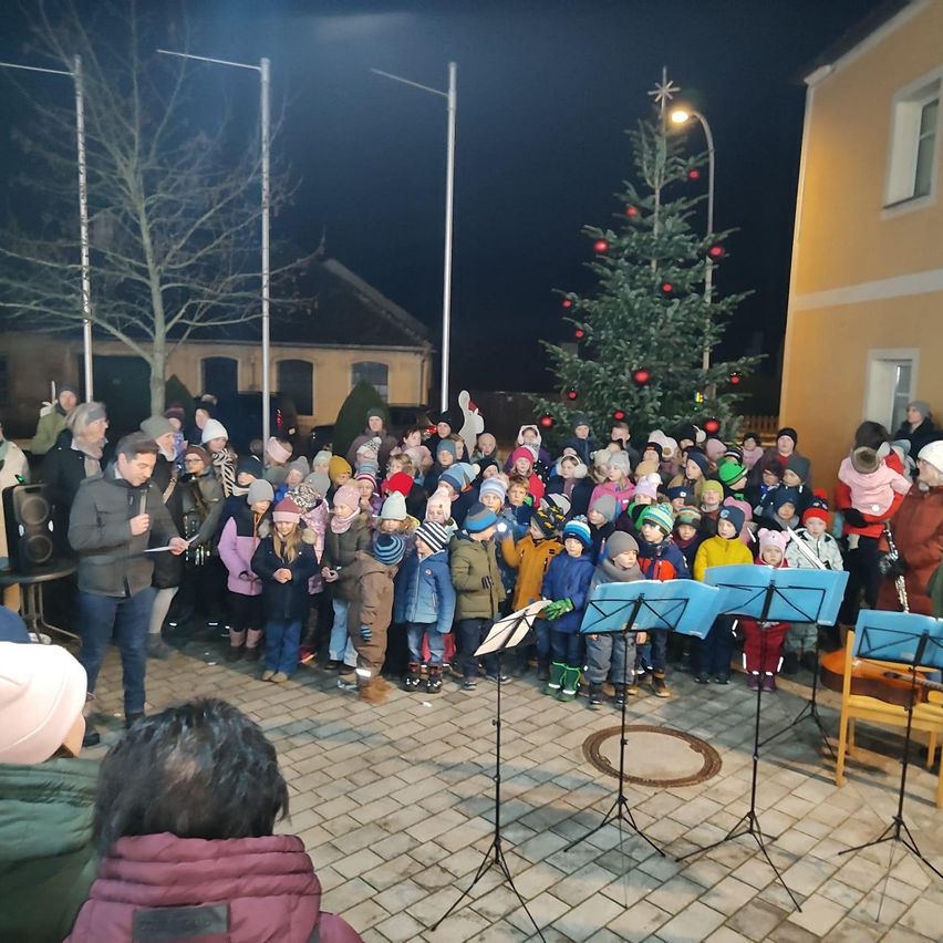 A crowd of children and adults gathered for a performance in front of a decorated Christmas tree at night, with musical instruments and street lights visible.