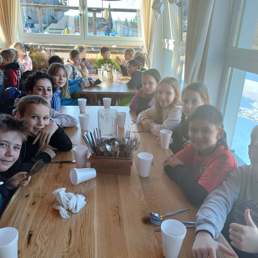 Eine Gruppe von Kindern sitzt an einem langen Holztisch und lächelt für die Kamera. Sie befinden sich in einem Restaurant mit großen Fenstern. Auf dem Tisch stehen Becher, Löffel und Gabeln.