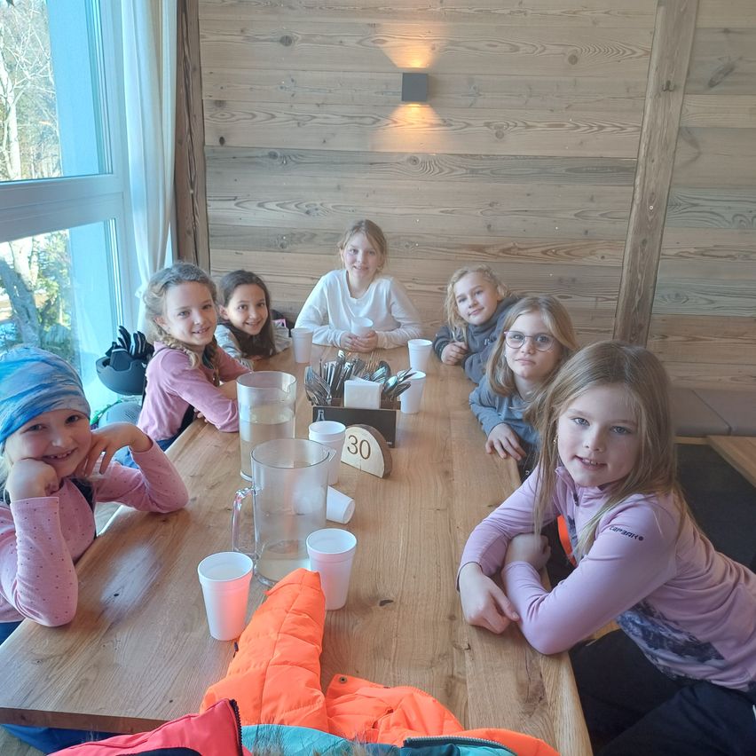 Eine Gruppe junger Mädchen und eine erwachsene Frau sitzen um einen Holztisch und lächeln für ein Foto. Sie tragen rosa Langarmshirts. Ein Glaskrug, Tassen und ein Schild mit der Aufschrift 30 stehen auf dem Tisch.