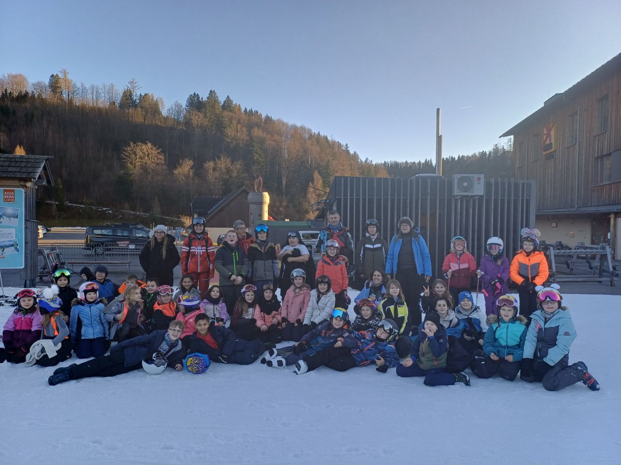 Eine Gruppe von Menschen, hauptsächlich Kinder, posiert für ein Foto auf einer verschneiten Oberfläche. Sie tragen Winterkleidung und einige halten Skistöcke. Hinter ihnen steht ein Gebäude mit einem Metallzaun.