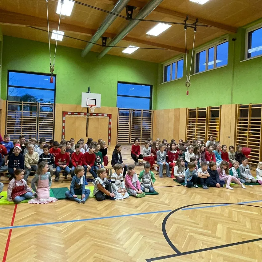 In einer Turnhalle sitzen Kinder in Reihen auf dem Holzboden und beobachten eine Aufführung oder Präsentation. Die Turnhalle hat grüne Wände, große Fenster und einen Basketballkorb im Hintergrund.