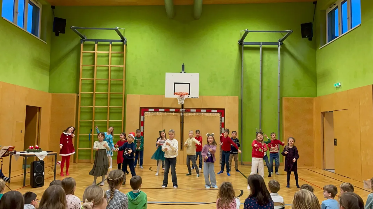 Eine Gruppe von Kindern tanzt auf einem Basketballplatz, einige tragen Feiertagspullover. Ein Basketballkorb ist an einer grünen Wand montiert, und in der Mitte befindet sich ein Netz. Ausrüstung ist an der Wand platziert.
