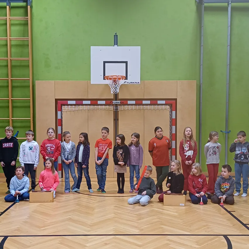 Eine Gruppe von Kindern versammelt sich in einer Turnhalle, trägt Winterkleidung und hält Musikinstrumente. Sie sitzen auf Holzkisten, während ein Basketballkorb im Hintergrund zu sehen ist.