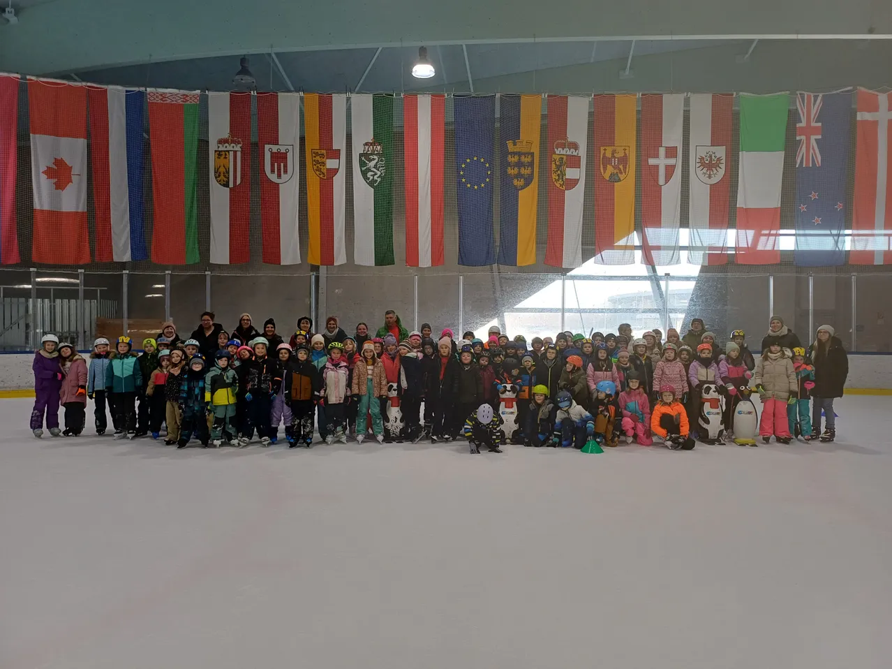 Eine Gruppe von Menschen in Winterkleidung posiert für ein Foto in einer überdachten Eisbahn mit verschiedenen Flaggen im Hintergrund.