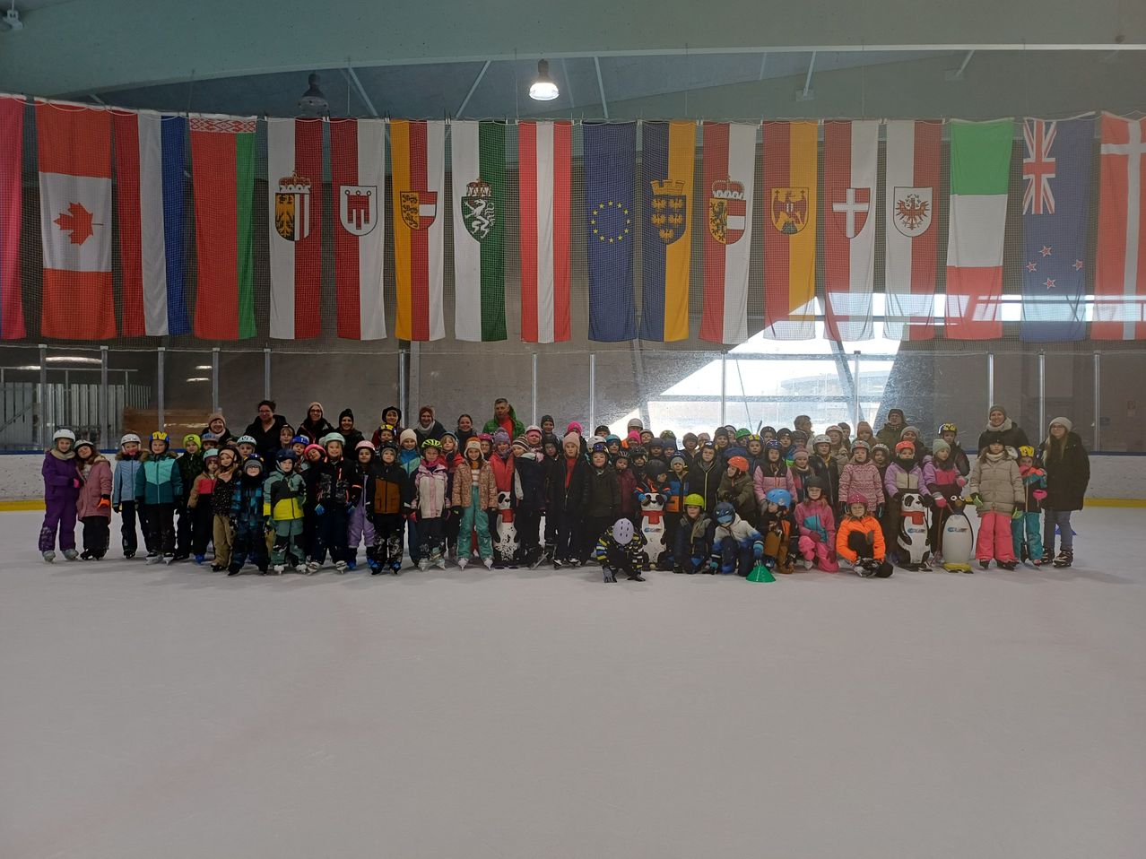 A group of people in winter gear pose for a photo in an indoor ice rink with various flags hanging in the background.