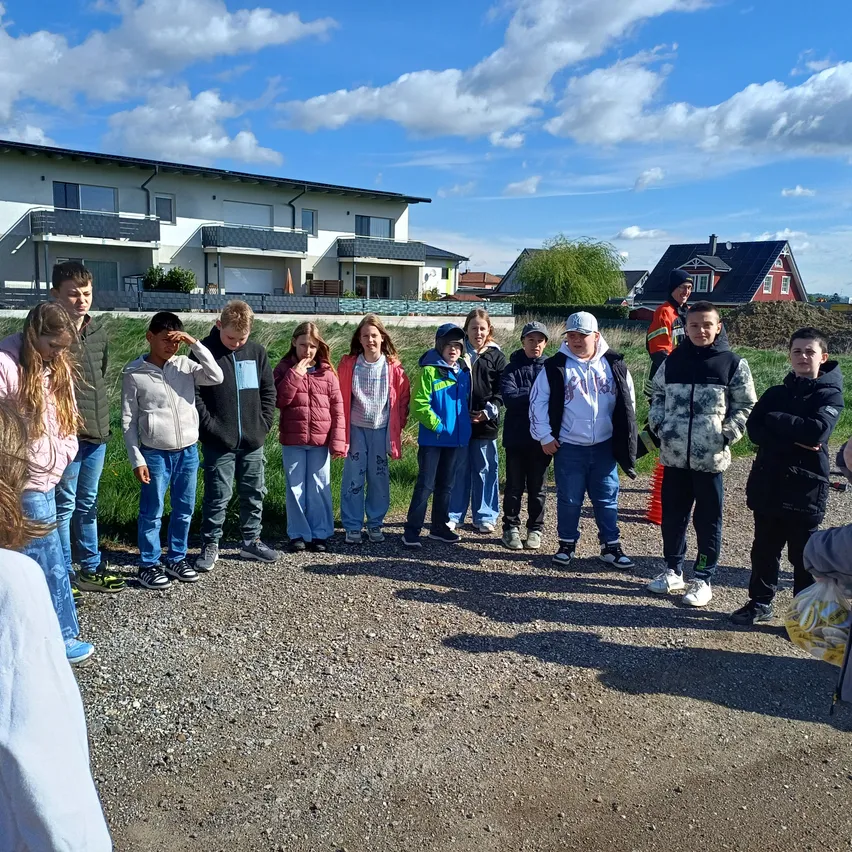 Eine Gruppe von Kindern und Erwachsenen steht vor einem Haus unter einem teilweise bewölkten Himmel, einige verdecken ihre Augen.
