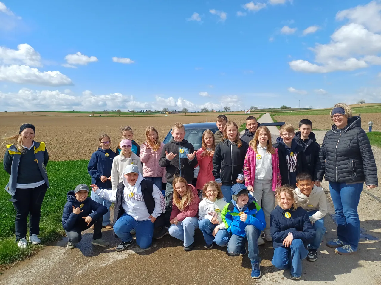 Eine Gruppe von Kindern und Jugendlichen posiert für ein Foto an einer Straße, mit einem Auto und einem Feld im Hintergrund.