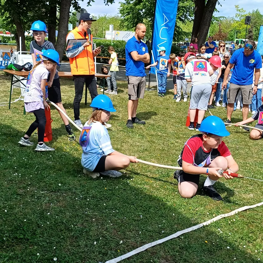 Kinder in blauen Schutzhelmen nehmen an einem Tauziehen auf einem Rasenfeld teil, während Zuschauer in der Nähe sind.