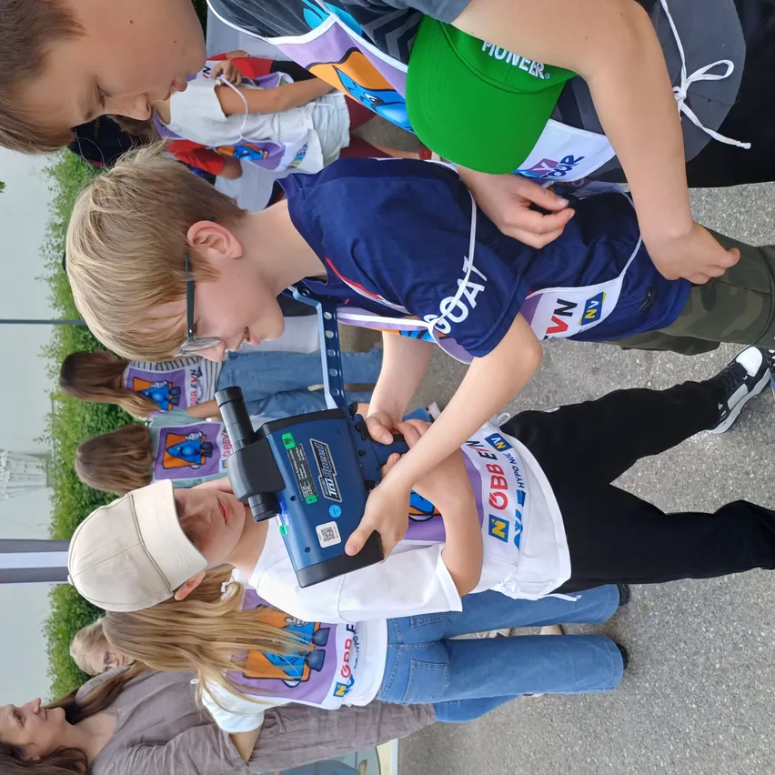 Gruppe von Kindern benutzt ein Gerät mit Bildschirm. Sie tragen verschiedene Hemden mit Logos. Ein Kind trägt eine Brille. Sie sind von anderen Kindern umgeben, einige mit Mützen.