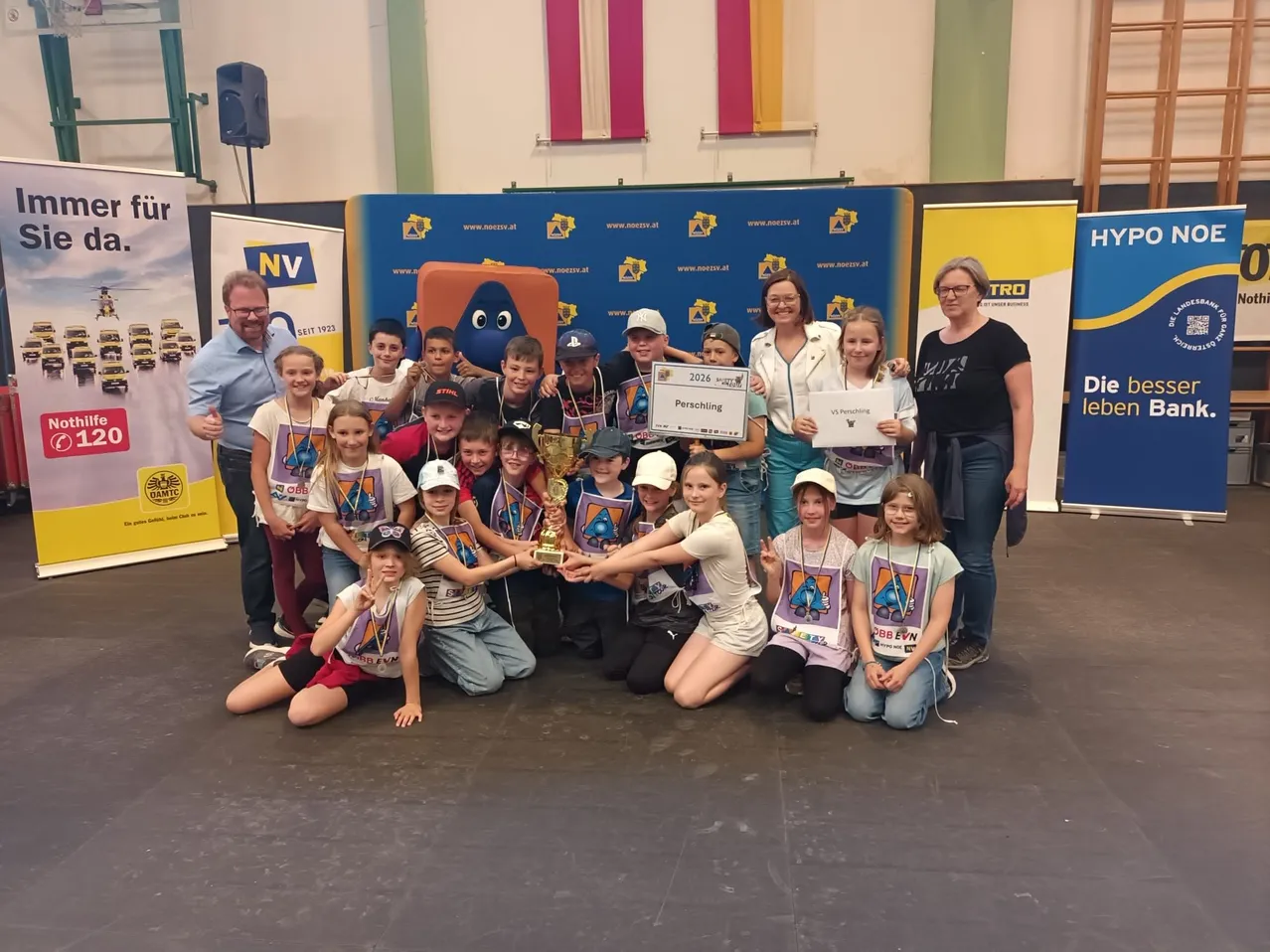 Eine Gruppe von Kindern und Erwachsenen posiert für ein Foto. Die Kinder halten einen Pokal, ein Zertifikat und eine Plakette. Sie tragen Shirts und Mützen. Hinter ihnen befinden sich Banner.