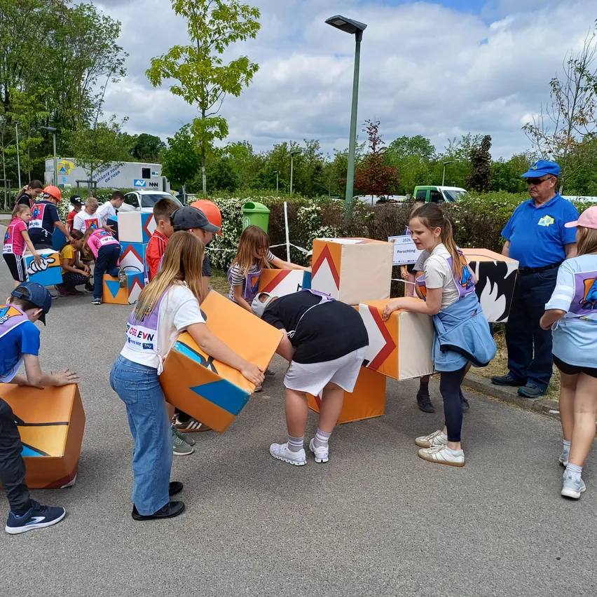 Eine Gruppe von Kindern und Erwachsenen versammelt sich um bunte Würfel, möglicherweise an einer Gemeinschaftsveranstaltung teilnehmend. Bäume und ein bewölkter Himmel im Hintergrund.