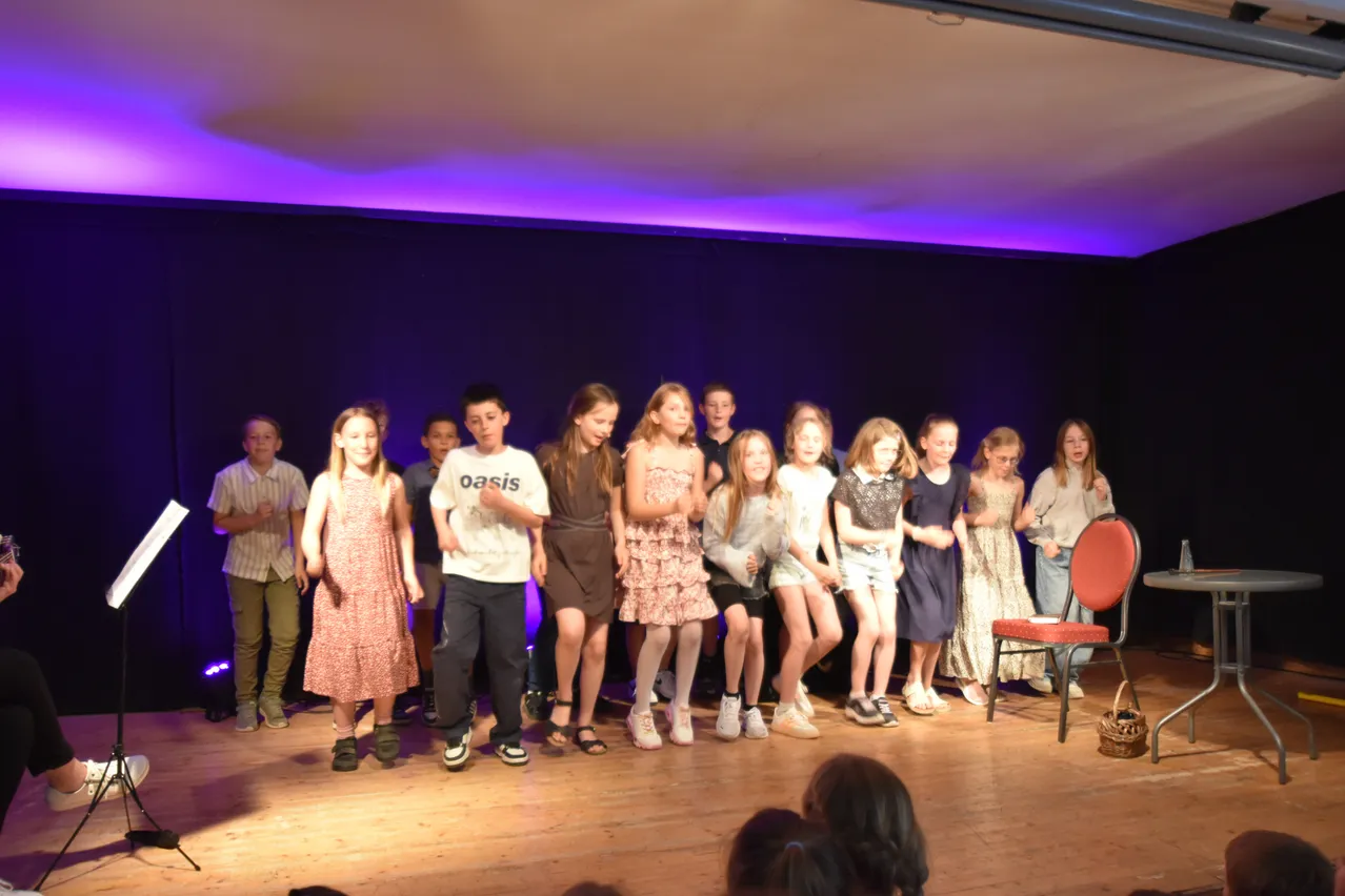 Eine Gruppe von Kindern in verschiedenen Outfits steht auf der Bühne, wahrscheinlich für eine Aufführung, unter violettem Licht.