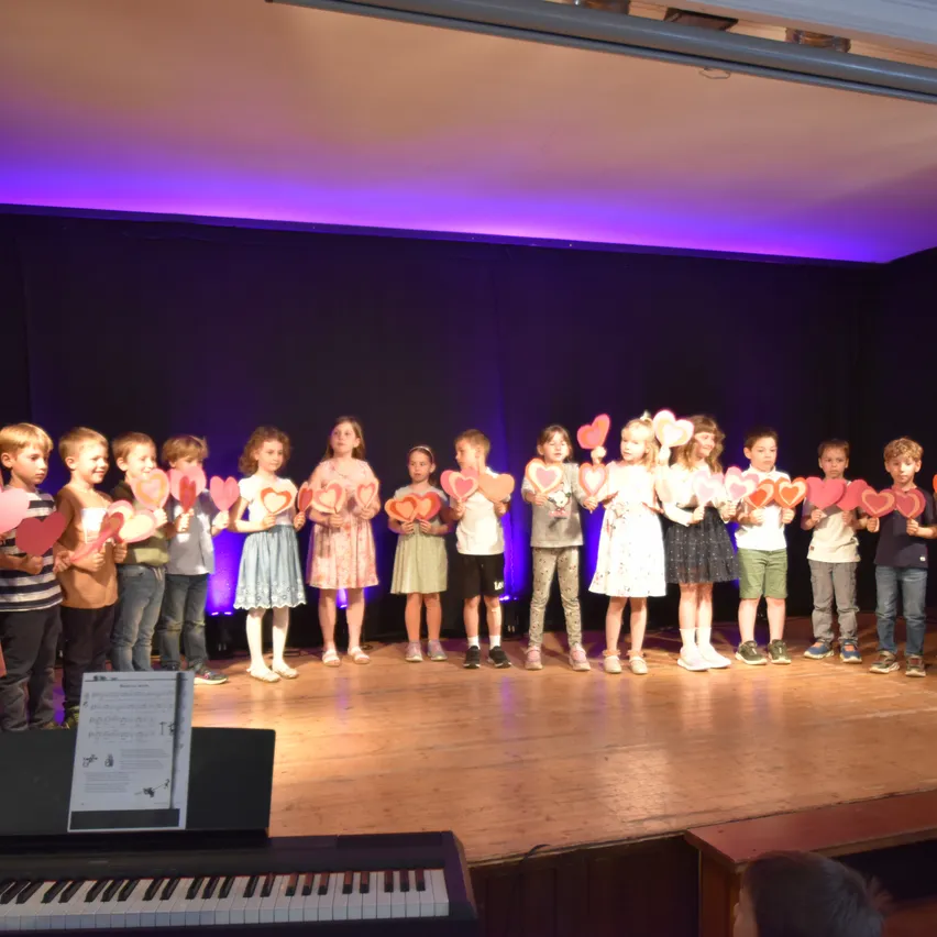 Eine Gruppe von Kindern auf der Bühne, die herzförmige Schilder halten, in verschiedenen Outfits gekleidet, mit einem Klavier davor und einem schwarzen Hintergrund.