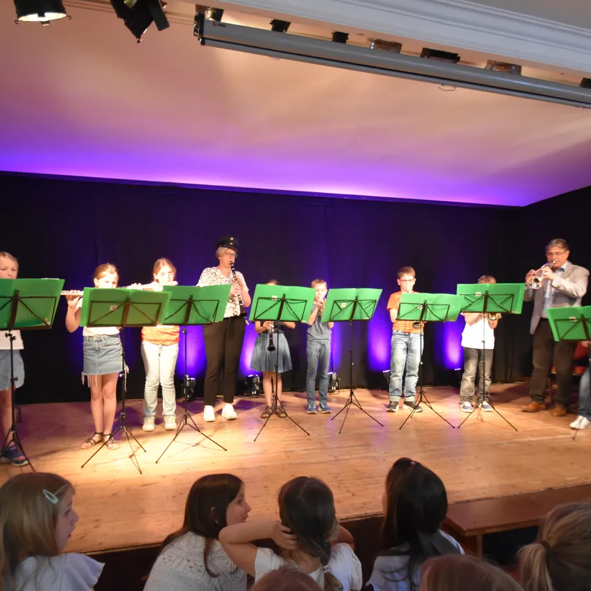 Eine Gruppe von Kindern und ein Erwachsener stehen auf der Bühne und spielen Musikinstrumente. Sie tragen alle passende Kleidung, einige halten Flöten und andere Trompeten. Die Bühne ist gut beleuchtet und das Publikum schaut zu.