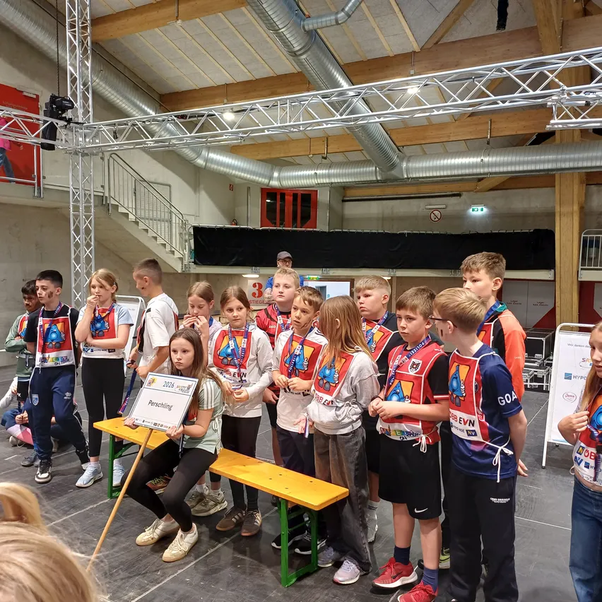 Eine Gruppe von Kindern steht auf einer Bühne, einige halten Banner. Ein Mädchen hält ein Schild mit '2024 VISION'. Sie tragen Uniformen und Medaillen. Hinter ihnen ist eine Bühne mit Treppen und einer Tür.