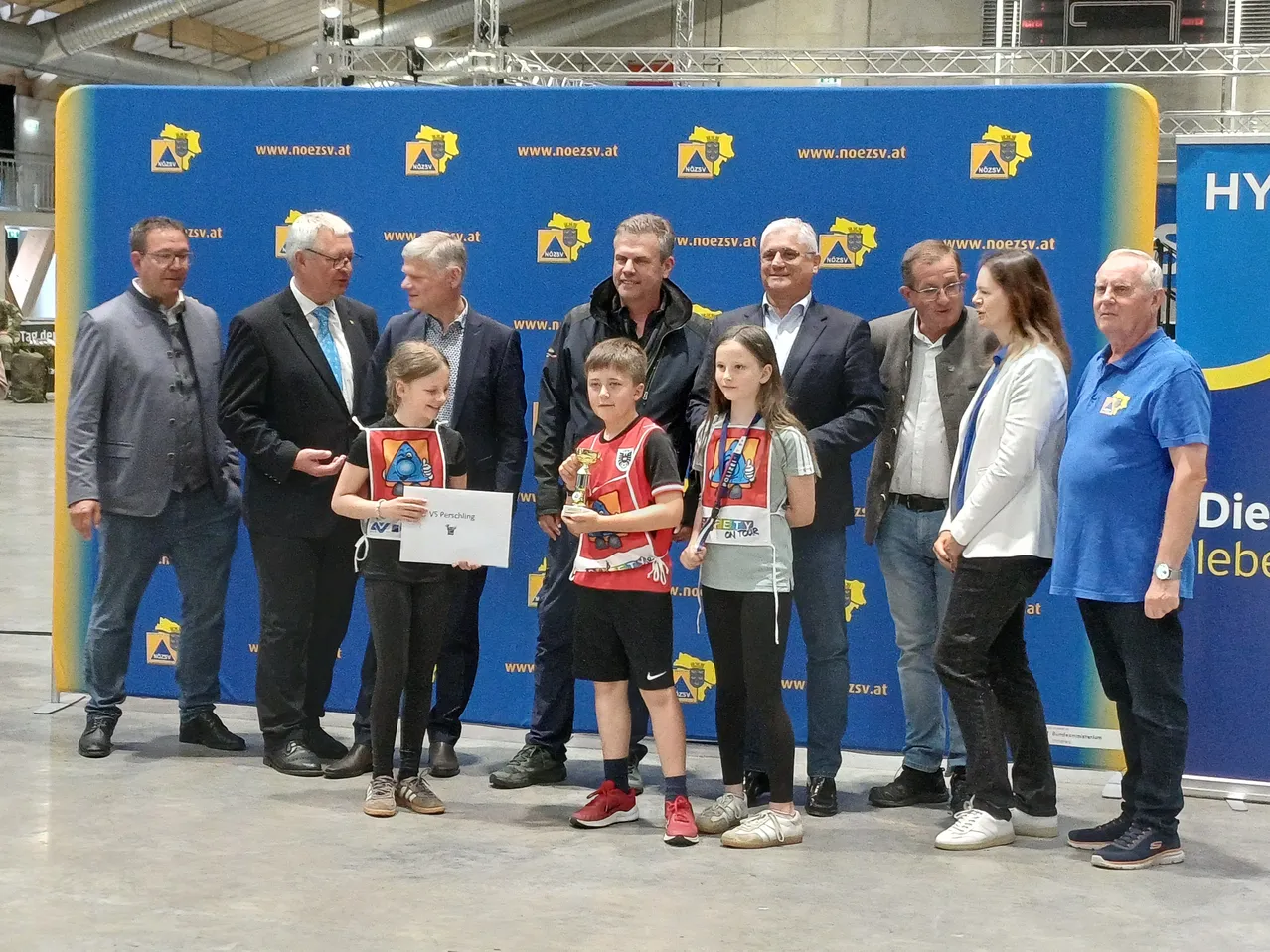 Eine Gruppe von Menschen steht vor einem blauen Hintergrund. Sie halten Auszeichnungen und sehen glücklich aus. Die Kinder tragen Sporttrikots und Turnschuhe. Die Erwachsenen sind in formeller Kleidung.