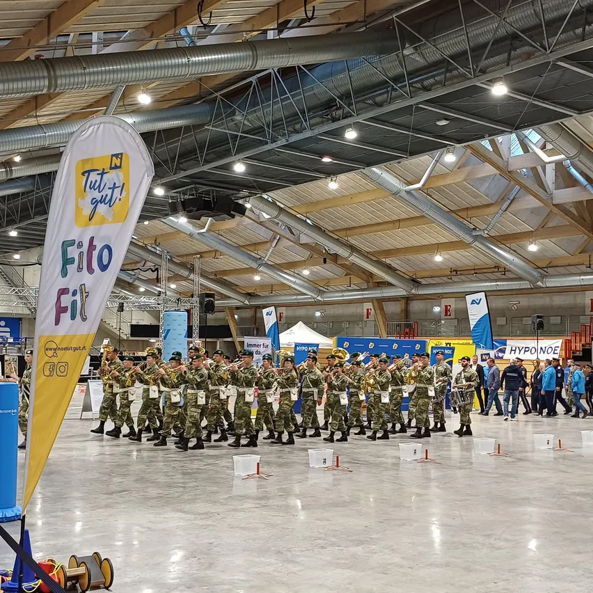 Eine Militärkapelle in grünen Uniformen steht in einer überdachten Arena und tritt auf. Zuschauer gehen durch die Arena, und Banner hängen von der Decke.