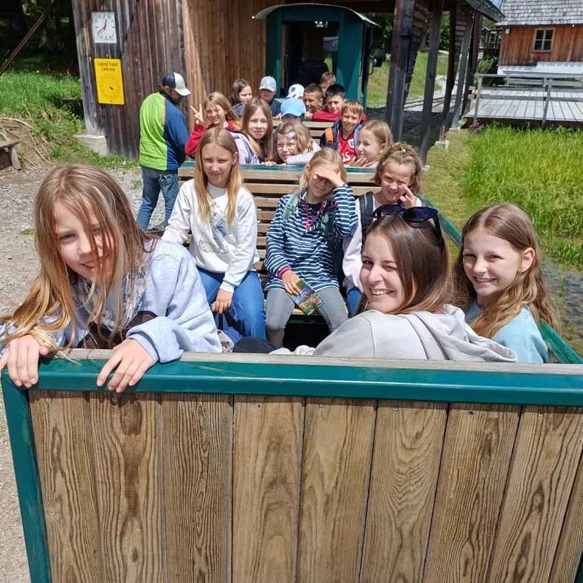 Eine Gruppe von Kindern und zwei Erwachsenen fährt in einem hölzernen Wagen auf einer Strecke, umgeben von Gras und Holzkonstruktionen.