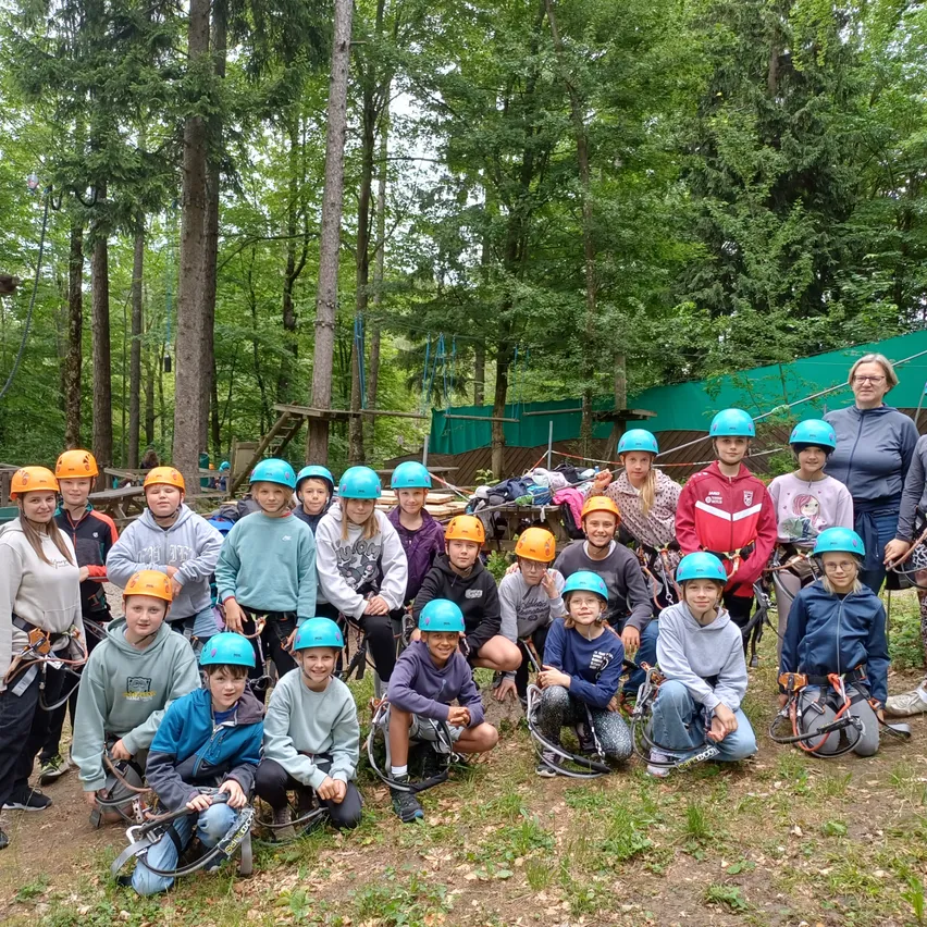 Eine Gruppe von Kindern mit orangen und blauen Helmen posiert für ein Foto im Wald. Sie stehen vor einer grünen Plane, mit Bäumen und einer Holzkonstruktion im Hintergrund.