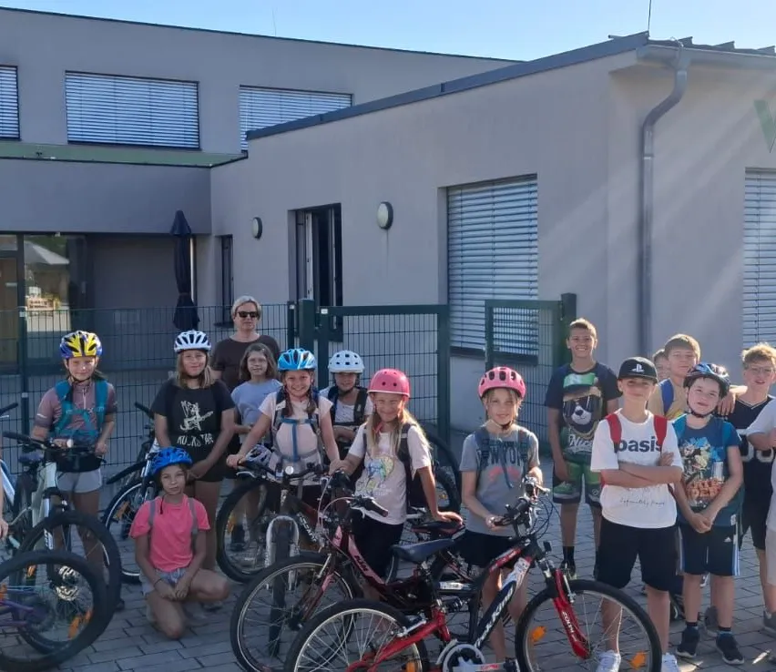 Eine Gruppe von Kindern in Helmen posiert mit ihren Fahrrädern vor einem Gebäude an einem sonnigen Tag.