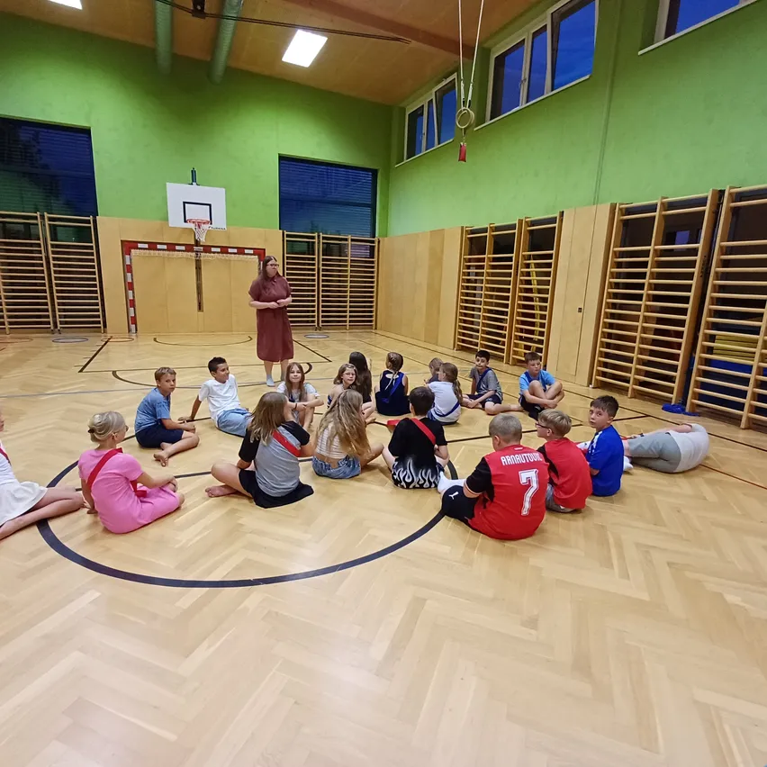In einer Turnhalle steht eine Frau vor einer Gruppe von Kindern, die im Kreis sitzen. Der Raum hat Holzwände, einen Basketballkorb und Fenster mit Jalousien.