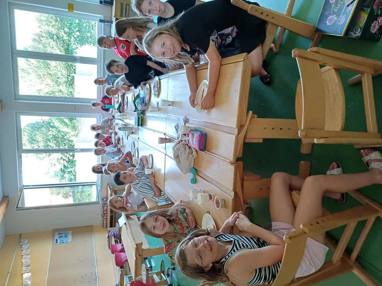 Eine Gruppe junger Kinder sitzt an Tischen in einem Klassenzimmer. Sie scheinen eine Mahlzeit oder einen Snack zu sich zu nehmen. Der Raum ist gut beleuchtet, wobei natürliches Licht durch große Fenster einfällt.