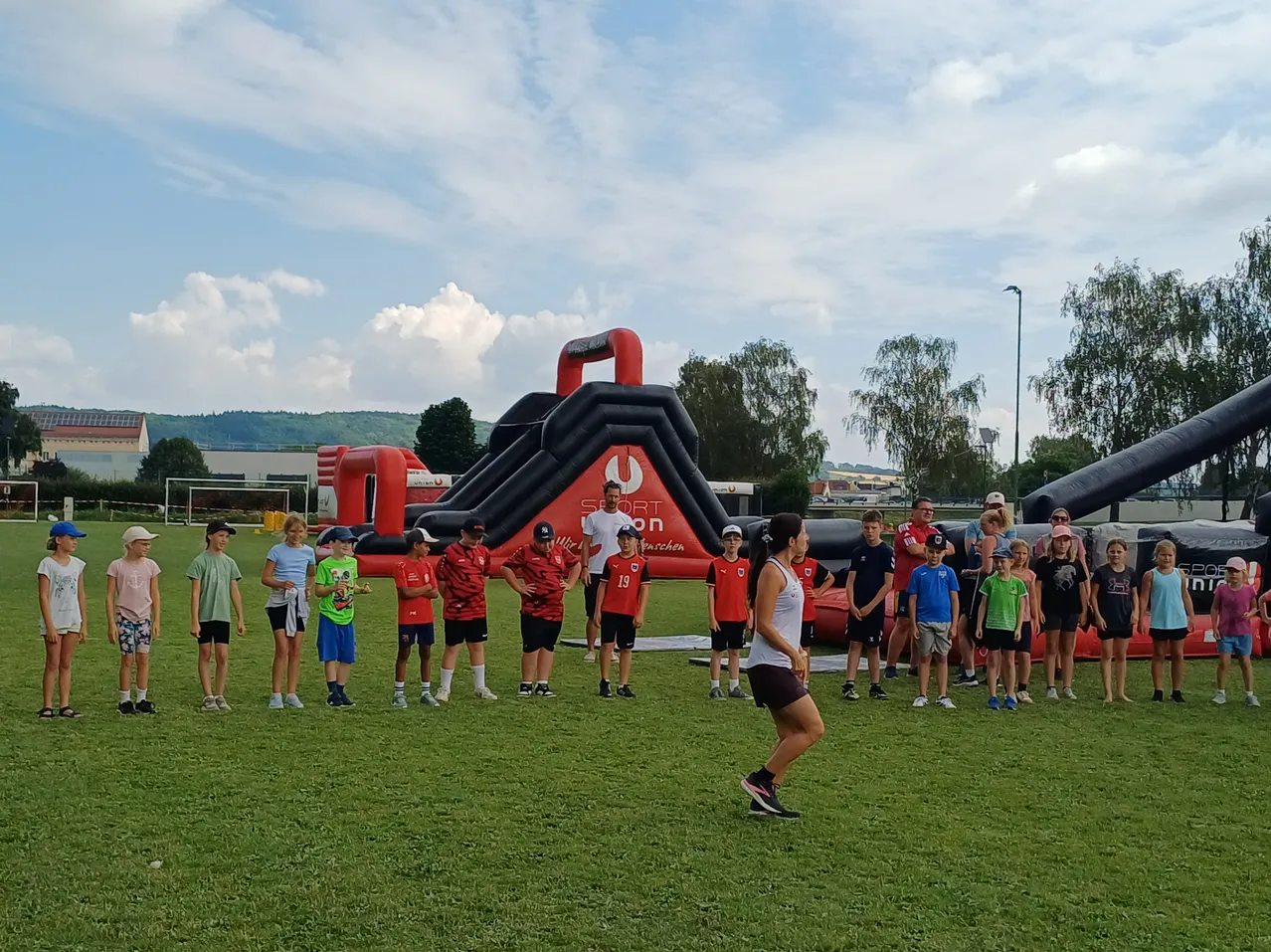 An einem sonnigen Tag stehen eine Gruppe von Kindern und Erwachsenen auf einem Feld mit einem großen roten aufblasbaren Hindernisparcours hinter ihnen. Einige beobachten eine Frau, die ein Spiel auf dem Feld spielt.