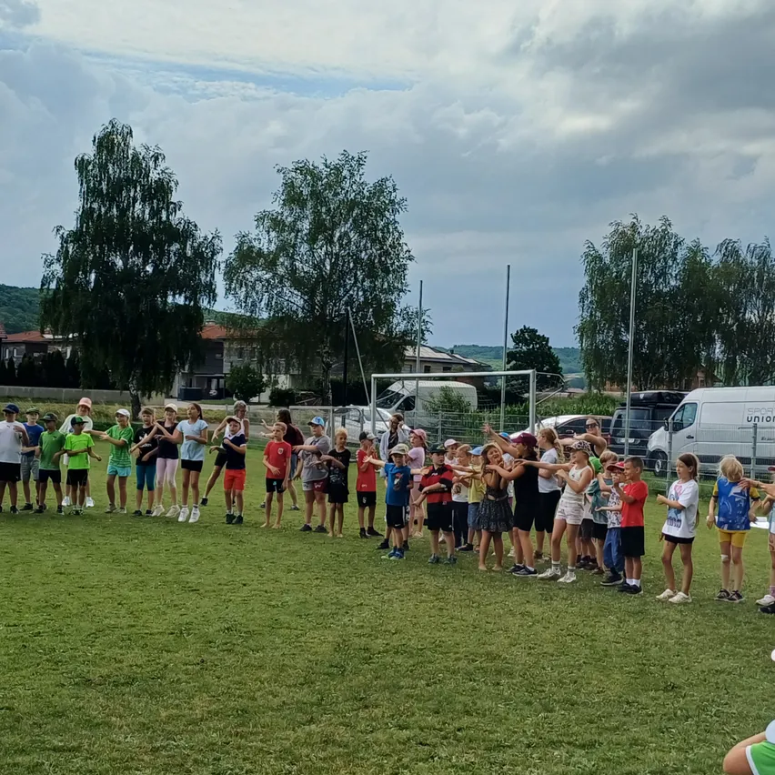 Eine Gruppe junger Menschen steht in einer Reihe auf einem Feld, einige halten Händchen, und ein Van ist in der Nähe geparkt.