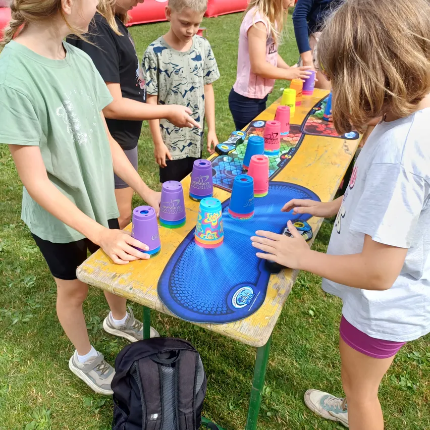 Mehrere Kinder spielen ein Spiel mit Bechern auf einem gelben Tisch im Freien. Sie stehen um den Tisch herum und versuchen, die Becher umzuwerfen.