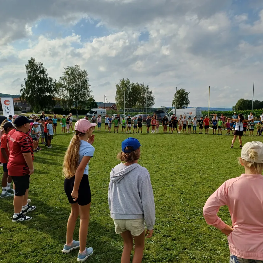 Kinder und Erwachsene versammeln sich auf einem Rasenfeld unter einem teilweise bewölkten Himmel. Einige tragen Mützen und Turnschuhe, während andere barfuß sind. Eine Person steht in der Nähe eines Torpfostens. Im Hintergrund sind Fahrzeuge geparkt.