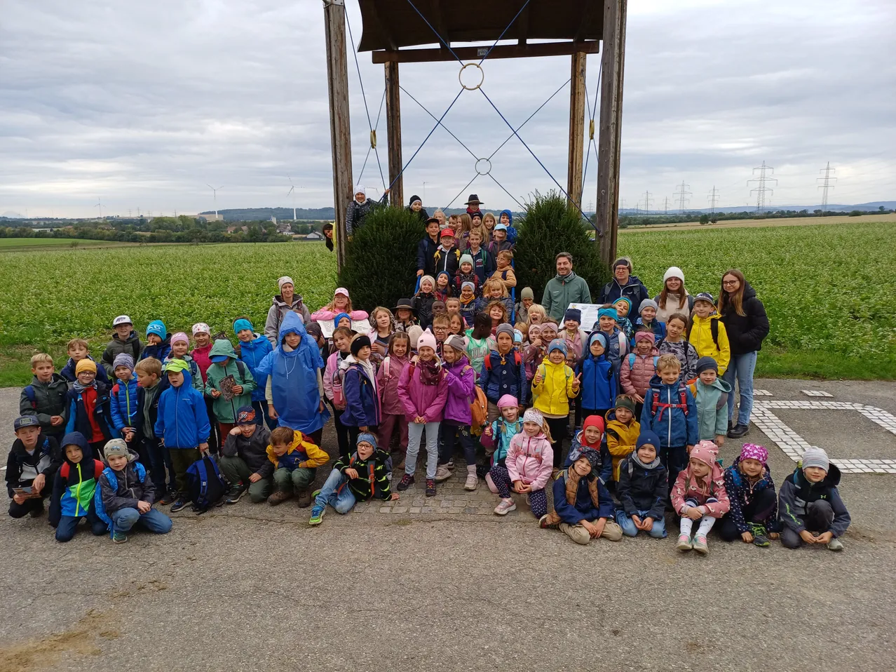 Eine große Gruppe von Kindern und Erwachsenen posiert für ein Foto auf einem Feld mit einer Holzstruktur im Hintergrund.