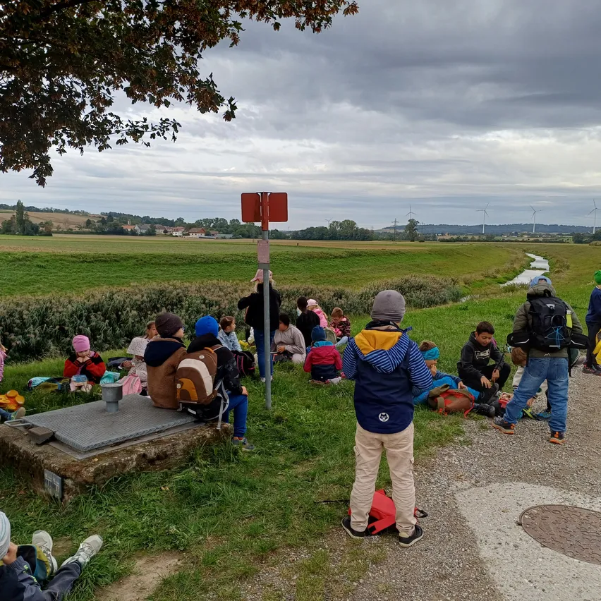 Eine Gruppe von Kindern und Erwachsenen sitzt auf der Wiese, einige tragen Mützen und Rucksäcke. Sie sind in der Nähe eines Schildes und eines kleinen Baches. Der Himmel ist bewölkt.