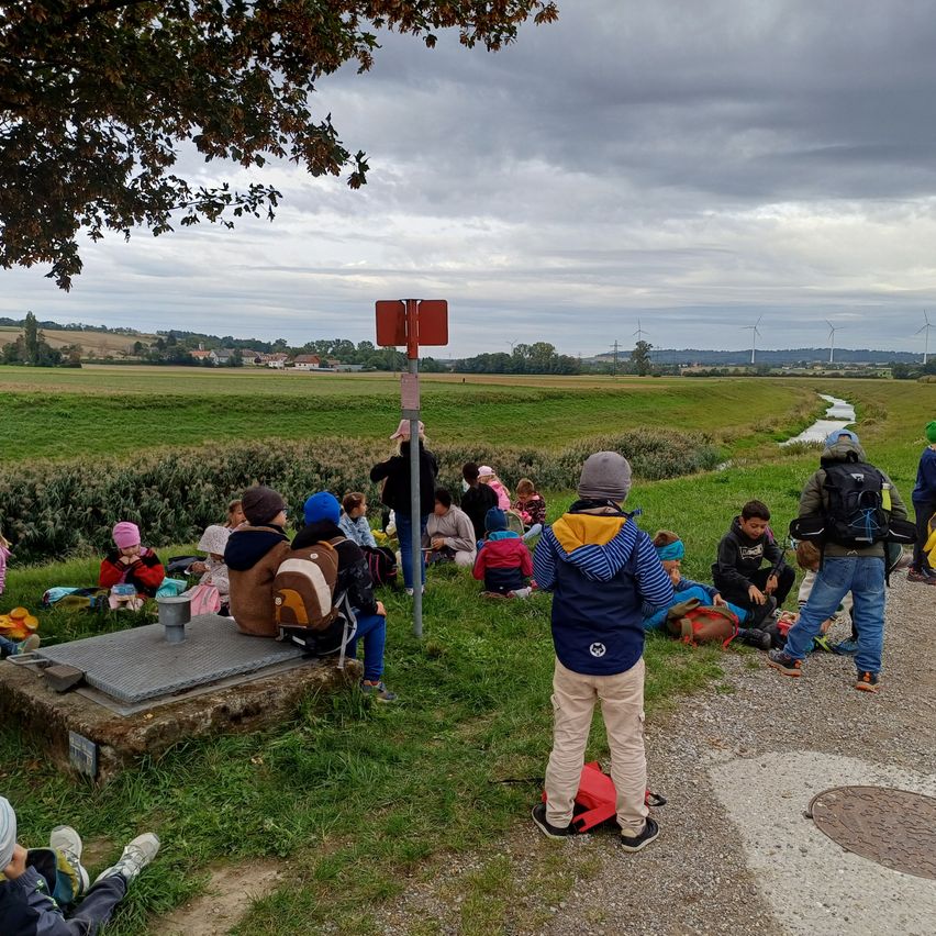 Eine Gruppe von Kindern und Erwachsenen sitzt auf der Wiese, einige tragen Mützen und Rucksäcke. Sie sind in der Nähe eines Schildes und eines kleinen Baches. Der Himmel ist bewölkt.