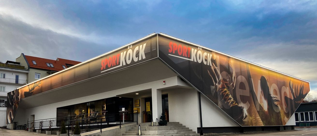 Ein Sportgeschäft mit einem großen Schild, das Sport Kock liest, zeigt ein Bild von Turnschuhen und Händen an seiner Außenwand, unter einem bewölkten Himmel.