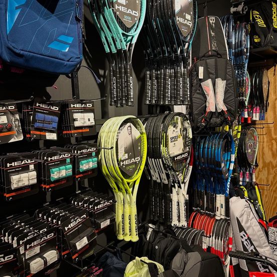 Ein Geschäft zeigt verschiedene Tennisschläger, darunter HEAD-Markenschläger, zusammen mit Sporttaschen und Zubehör an einer Wand an.