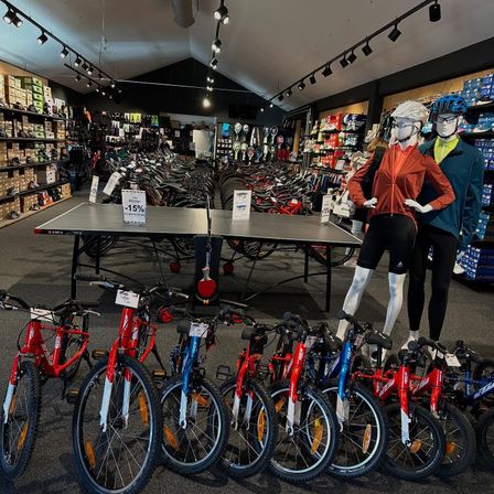 Fahrräder in verschiedenen Farben werden in einem Geschäft mit Mannequins in Fahrradbekleidung ausgestellt. Im Hintergrund befindet sich ein Tischtennistisch mit einem Rabattschild.