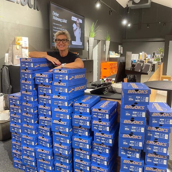 Eine lächelnde Frau steht hinter einem großen Stapel blauer Brooks-Schuhkartons in einem Einzelhandelsgeschäft. Hinter ihr befindet sich ein großer Bildschirm mit einer Werbebotschaft.