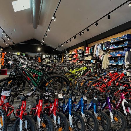 Ein Fahrradgeschäft mit einer großen Auswahl an Fahrrädern, einschließlich Kinderfahrrädern, in Reihen angeordnet. Verschiedene Farben und Größen sind ausgestellt. Im Hintergrund befinden sich Regale mit Schuhen und Kleidung.