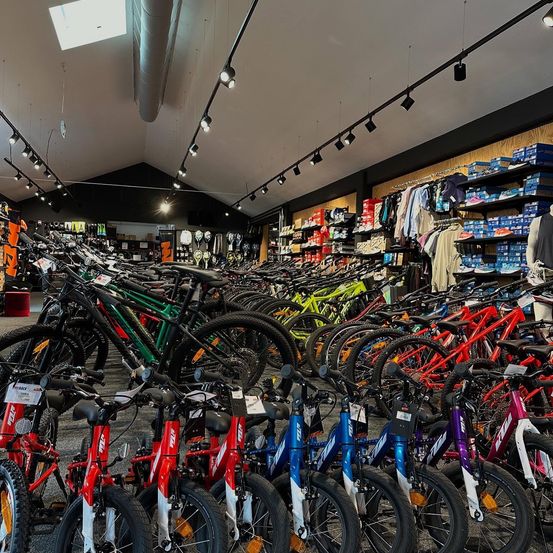 Ein Fahrradgeschäft mit einer großen Auswahl an Fahrrädern, einschließlich Kinderfahrrädern, in Reihen angeordnet. Verschiedene Farben und Größen sind ausgestellt. Im Hintergrund befinden sich Regale mit Schuhen und Kleidung.