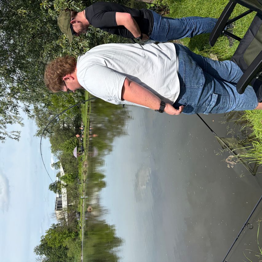 Two men fishing by a lake, one in white shirt and the other in black, with trees and a building in the background.