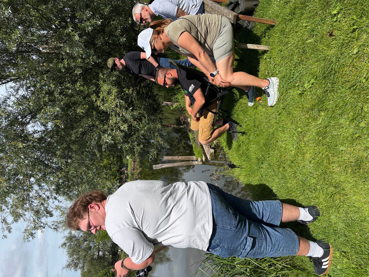 A group of people are on a grassy field, two are fishing by a river. One is bending over, another is lying on the grass. Two are standing near a tree, one holding a camera.