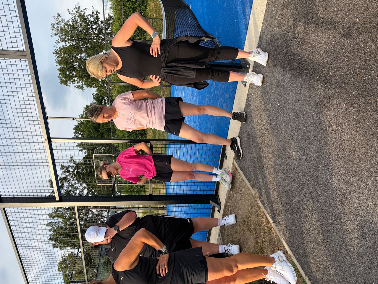 Four people in athletic wear stand on a court. One woman leans on a fence, while another looks down. The two men wear watches and caps. Trees and a fence are in the background.