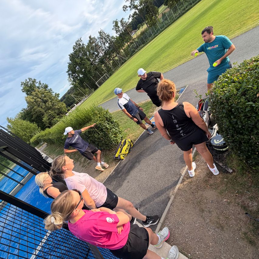 A group of people in athletic wear are on a tennis court. One person holds tennis balls, and another is bent over, possibly talking. A tennis racket rests on the ground.