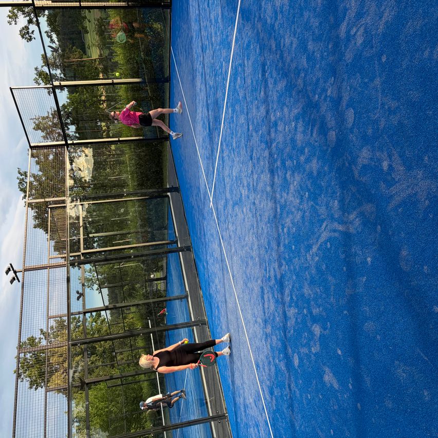 Two players are playing tennis on a blue court, one in a pink shirt and the other in black. The court is enclosed by a fence.